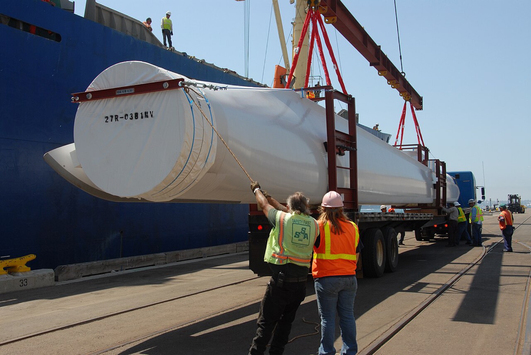 Workers unloading a large wind turbine blade from a ship to a truck